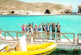 A group of students waving next to a boat at the Blue Lagoon, Comino